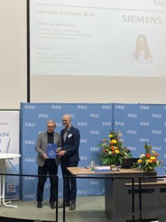 Two men standing on a podium. The one on the left is receiving a certificate.